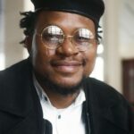 Confident man in glasses with a stylish hat smiling indoors.